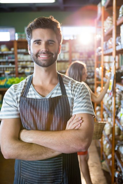 Male staff member standing with arms crossed in the organic section of a supermarket, smiling confidently. Ideal for use in advertisements for grocery stores, organic food markets, or small businesses. Highlights customer service and friendly staff in a retail environment.
