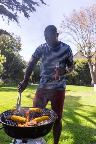 African American man grilling burgers and corn in sunny backyard holding drink bottle and tongs