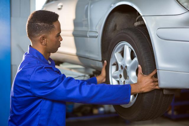 Mechanic changing a car tire in a repair garage, wearing blue overalls, focuses on ensuring the wheel is properly aligned and secure. Suitable for use in marketing materials for auto repair shops, car maintenance guides, vehicle service advertisements, and educational pieces on automotive care.