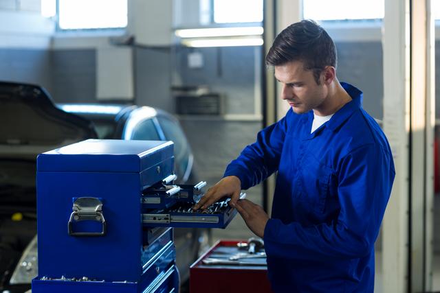 Mechanic in blue uniform checking tools in a toolbox at an auto repair garage. Ideal for use in articles or advertisements related to automotive repair services, mechanic training programs, or tool and equipment suppliers.