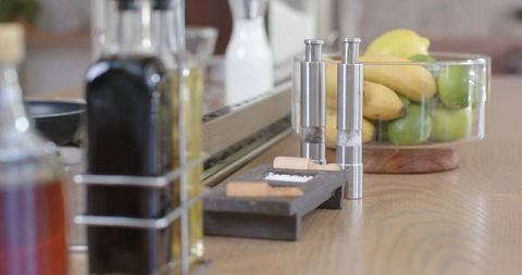 Stainless-steel spice grinders standing on wooden countertop with salt dish and fruit bowl