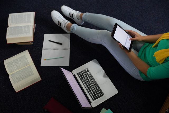 Asian female student sitting on the floor in a library, using a tablet and laptop for studying. Books and a notebook are scattered around her. Ideal for educational content, online learning platforms, and technology in education themes.