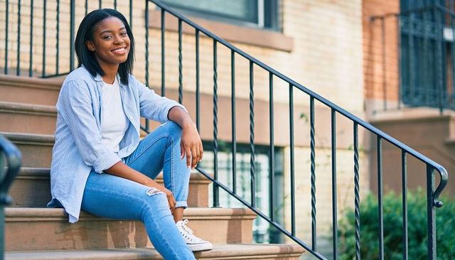 Young woman sitting on brownstone steps smiling in casual striped shirt and ripped jeans