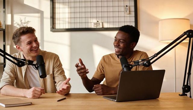 Podcast hosts laughing and collaborating while recording in casual home studio with laptop