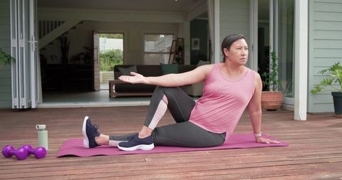 Mature woman doing seated spinal twist on pink mat on wooden deck during home workout