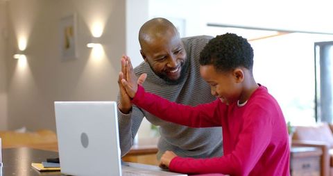 African american father and son sharing high-five while learning on laptop in kitchen