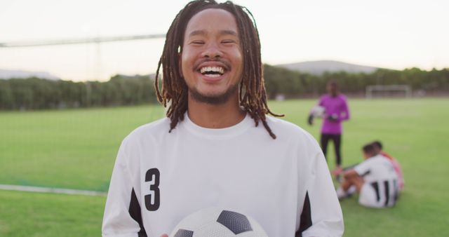 Young soccer player wearing white jersey with number 3, smiling and holding a ball on a soccer field. Teammates in the background. Ideal for sports, teamwork, and youth-related themes.