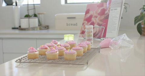 Pink frosted vanilla cupcakes cooling on rack in bright minimalist kitchen with piping bag