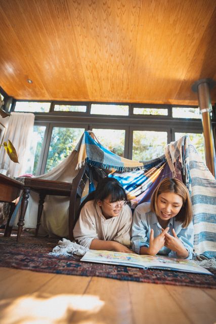 Happy asian mother and daughter lying on floor in blanket camp reading book together and smiling. at home in isolation during quarantine lockdown.