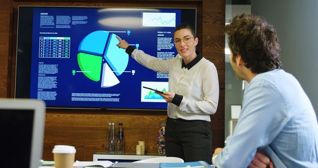 Businesswoman is presenting data on a large screen to a colleague in a modern office setting. She uses a tablet and points at the chart with confidence. Ideal for depicting concepts like teamwork, business meetings, data analysis, and modern professional environments. Suitable for business presentations, corporate training materials, and articles about workplace collaboration.