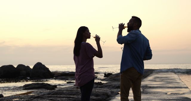 Biracial couple enjoying drinks by sea at sunset. She has long dark hair, he's wearing blue shirt, both looking relaxed