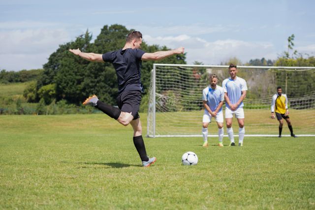 Football player taking a penalty shot on a grassy field with teammates and goalkeeper in the background. Ideal for use in sports-related content, advertisements, and articles about soccer, teamwork, and athletic skills.