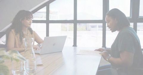Women collaborating at office conference table reviewing documents and working on laptop