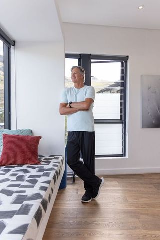Mature man standing by window in modern sunlit room wearing smartwatch and leaning on daybed