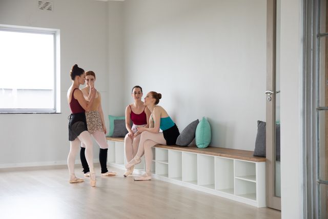 Group of Caucasian female attractive ballet dancers warming up and practicing in a bright ballet studio, standing and sitting on a bench. Preparing for a class and talking.