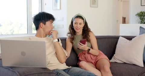 Young couple planning weekend trip on couch using laptop and smartphone in modern living room