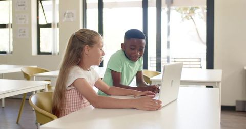 Diverse students collaborating on laptop for digital learning in bright modern classroom