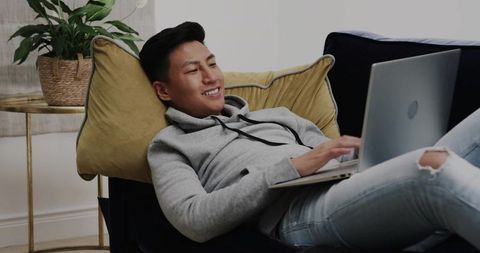 Reclining asian man typing on laptop on sofa with mustard pillow for remote work and relaxation