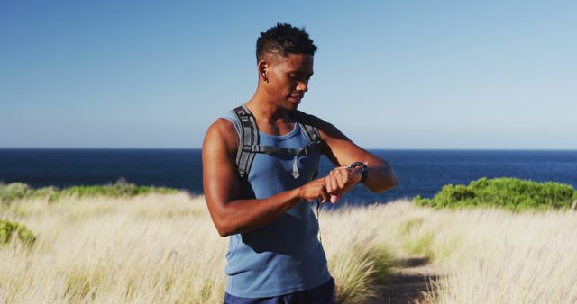 African american man cross country running using smartwatch in countryside by the coast. fitness training and healthy outdoor lifestyle.