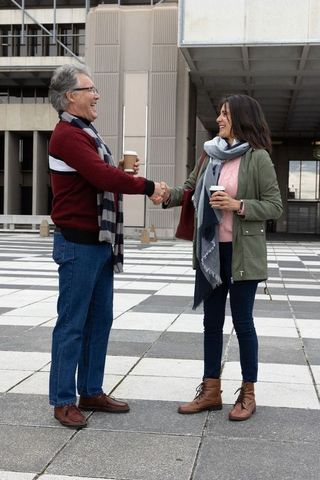 Mature couple shaking hands while holding coffee cups in urban plaza