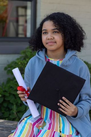 Female teen celebrating graduation holding mortarboard and diploma on bench outdoors campus