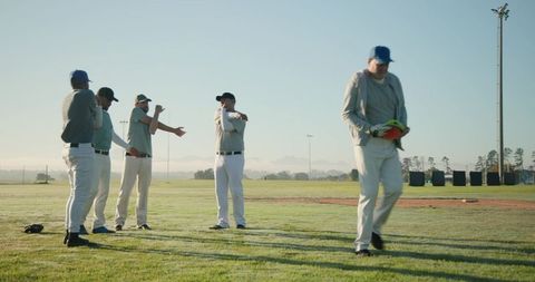 Baseball Team Warming Up at Sunrise on Grass Field with Coach Walking Forward Holding Gear