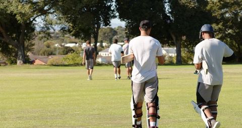 Cricket team walking across sunny park field carrying bats and wearing pads