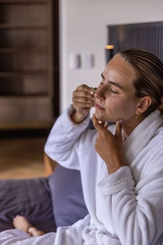 Woman using jade facial roller while relaxing in plush white robe at home spa