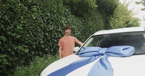 Young man receiving new car gift with giant blue bow in suburban driveway