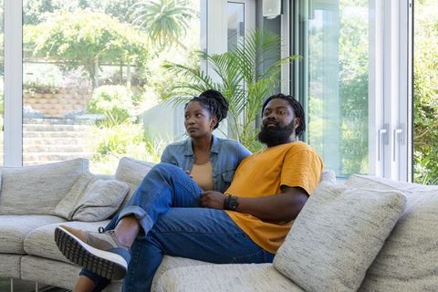 African couple relaxing on modern light-gray sectional sofa near sunlit glass doors
