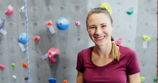 Young woman smiling while standing in an indoor climbing gym. She is wearing a maroon shirt with a simple ponytail hairstyle. The climbing wall is equipped with colorful holds for rock climbing activities, indicating a vibrant and engaging environment. Ideal for illustrating concepts of fitness, active lifestyle, climbing activities, personal achievements, and motivation.