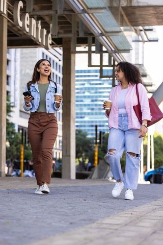 Two african american women walking downtown holding coffee cups and phone, laughing together