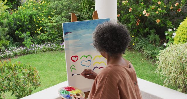 Elderly woman enjoying a creative painting session outdoors in a beautiful garden, illustrating colorful hearts on canvas. Ideal for topics related to senior hobbies, art therapy, creative aging, mental well-being, and leisure activities in nature.