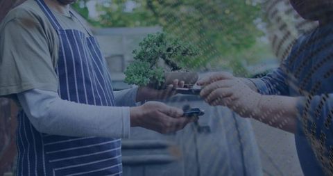 Vendor handing bonsai while customer tapping phone for contactless market transaction