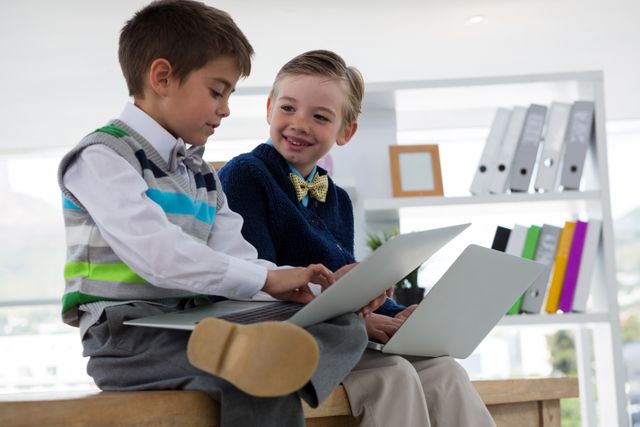 Two young boys dressed in formal attire are sitting on a table in an office environment, using laptops. One boy is typing while the other looks on with interest. The background includes office supplies and shelves, suggesting a professional setting. This image can be used to illustrate concepts of young entrepreneurship, early education in business, teamwork, and the integration of technology in learning.