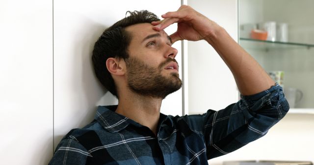 Young man in a modern kitchen, displaying stress and contemplation with hand on forehead. Useful for depicting stress, anxiety, mental health, and emotional challenges in daily life.