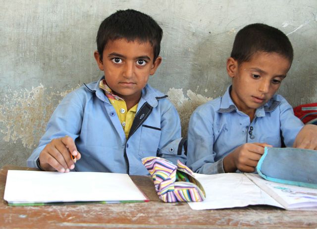 Two young boys are sitting at a wooden desk in a classroom, dressed in school uniforms. One boy is writing with a pencil, capturing a determined look. The other boy is concentrating on his workbook and school supplies, intensely focused. The image highlights themes of education, learning, and childhood. This would be perfect for educational materials, blog articles about childhood education, school promotions, or charitable organizations focusing on education.