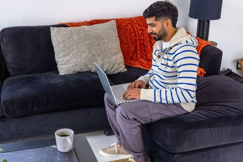 Asian man typing on laptop on velvet sofa at home, cozy living room with coffee and throw