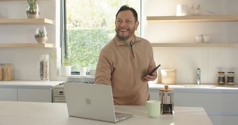 Smiling man using laptop and smartphone in modern kitchen