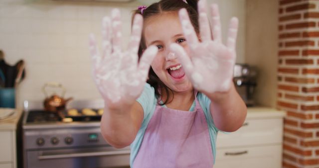 Smiling young girl baking in kitchen, showing her hands covered in flour. Ideal for use in family and culinary blogs, advertisements for kitchenware or children's activities, and promotional material for baking classes.