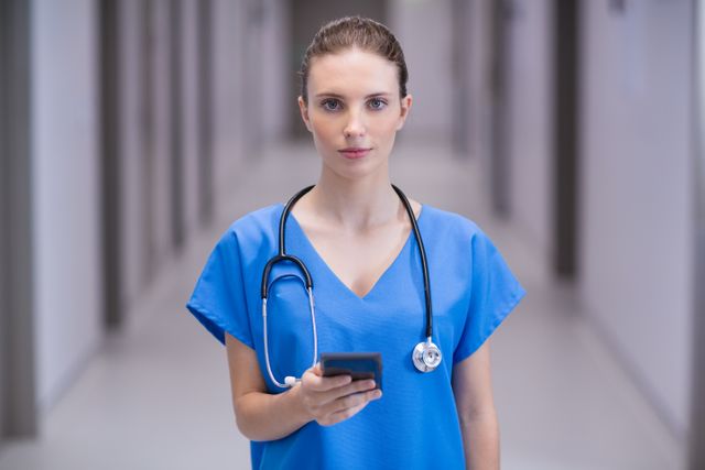 Female doctor in medical uniform standing in hospital corridor and using mobile phone. Ideal for illustrating modern healthcare, medical technology, and professional healthcare services. Useful for medical blogs, healthcare websites, and hospital promotional materials.
