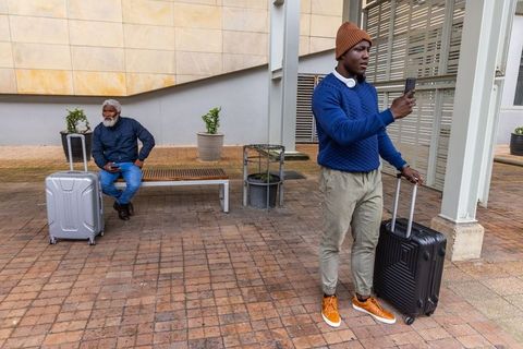 Young man checking phone while waiting with luggage at urban transit plaza near bench
