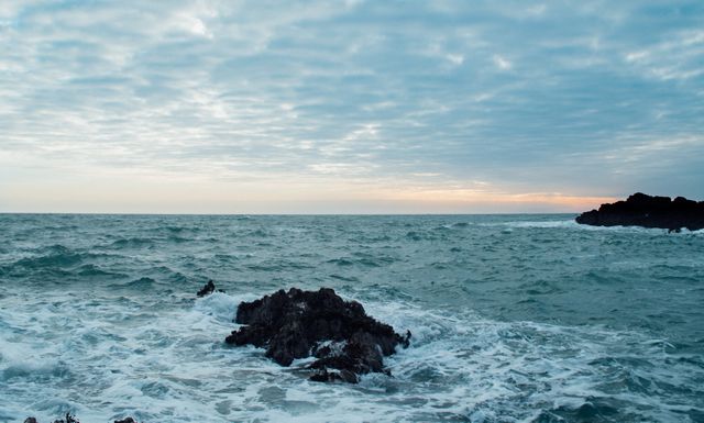 Dramatic sea waves crashing against rocky shore at dusk, with a cloudy sky over the horizon. This scene captures the intensity and beauty of coastal landscapes. Ideal for backgrounds, environmental concepts, and content related to nature, travel, and weather.