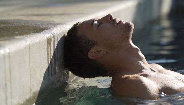 Sunlit bare-chested man resting in pool leaning on concrete coping, serene profile closeup
