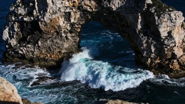 Crashing waves through limestone arch on rugged coastal cliff, dramatic foamy surf