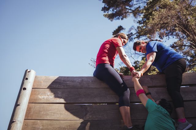 Woman being assisted by her teammates to climb a wooden wall during obstacle course training at boot camp