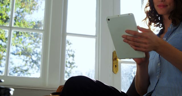 A young Caucasian woman is casually using a tablet near a window with natural light streaming in, with copy space. Her relaxed posture and the bright setting suggest a comfortable, leisurely activity at home or a casual workspace.