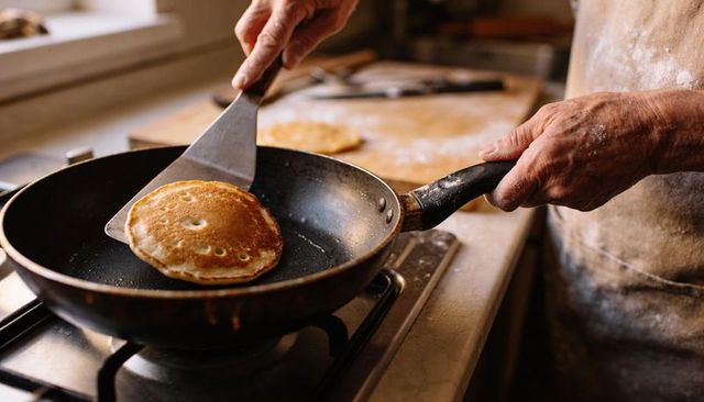 Senior cook flipping golden pancake on nonstick pan at gas stove in sunlit rustic kitchen