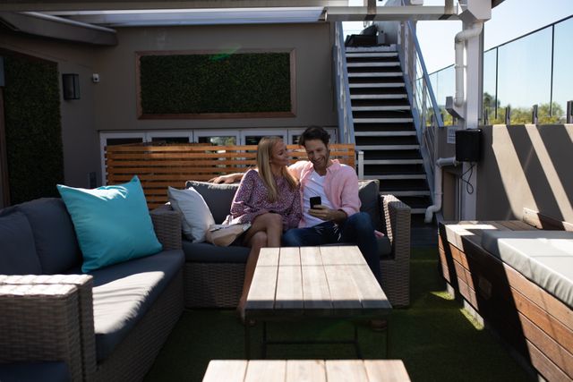 Caucasian couple enjoying leisure time on an outdoor hotel terrace. They are sitting together on a sofa, smiling and talking while the man uses a smartphone. Ideal for themes related to vacation, relaxation, modern lifestyle, technology, and bonding moments.