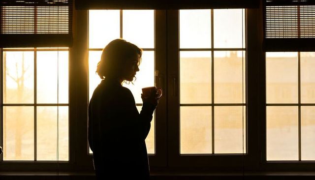 Silhouette woman standing holding mug at golden hour by paned window blinds